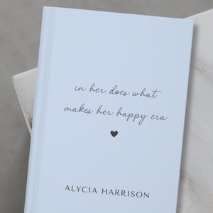 May include: A light blue journal with the text "in her does what makes her happy era" and a small black heart. The author's name, "ALYCIA HARRISON", is printed at the bottom of the cover.