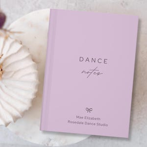 May include: A lavender-colored notebook with the words "DANCE notes" printed on the cover. Below the title is a bow graphic and the text "Mae Elizabeth Rosedale Dance Studio". The notebook is resting on a white marble surface.