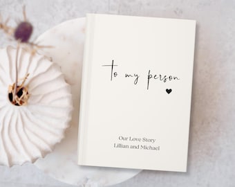 May include: White journal with the text "To my person" in black script with a small black heart. The text "Our Love Story" is below with the names "Lillian and Michael" underneath.