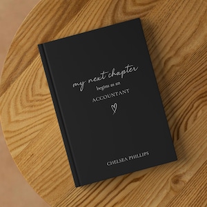 May include: A black journal with white script reading "my next chapter begins as an ACCOUNTANT". A small heart symbol is below the word "ACCOUNTANT". The name "CHELSEA PHILLIPS" is printed at the bottom of the journal.