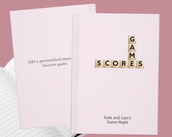 Puede incluir: Un cuaderno rosa con una cubierta blanca y páginas rayadas. La cubierta tiene las palabras "SCORES" y "GAMES" deletreadas con fichas de Scrabble. El texto "Kate and Cam's Game Night" está impreso debajo de las fichas.