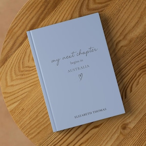 May include: A pale blue journal with the words "my next chapter begins in AUSTRALIA" and a heart symbol. The name "ELIZABETH THOMAS" is printed at the bottom. The journal rests on a wooden table.