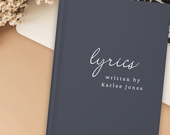 May include: A navy blue notebook with the word "lyrics" in elegant white script. Below, the text reads "written by Karlee Jones." The notebook is on a beige surface next to a laptop keyboard and dried flowers.