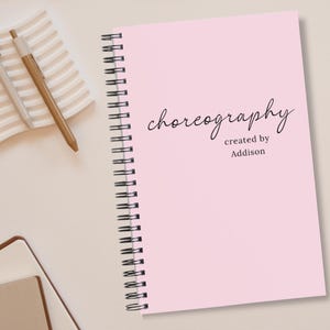May include: A pink spiral-bound notebook with the word "choreography" in cursive script. The text "created by Addison" is below. Two pens rest on a beige wavy tray, and a partially visible notebook is in the lower left corner.