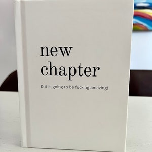 May include: White notebook with the text "new chapter & it is going to be fucking amazing!" printed on the front.