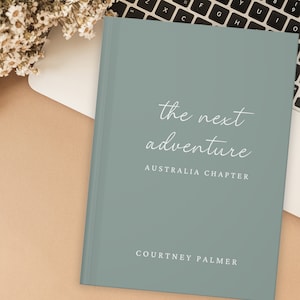 May include: A teal blue hardcover journal with the text "the next adventure" and "AUSTRALIA CHAPTER" in white. The bottom of the journal has the text "COURTNEY PALMER" in white.