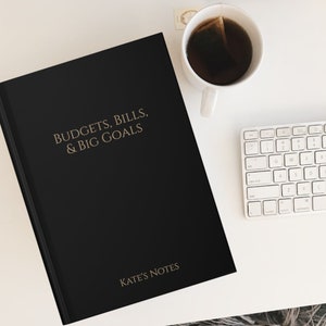 May include: A black notebook with the text "BUDGETS, BILLS, & BIG GOALS" and "KATE'S NOTES" in gold lettering. The notebook is on a white surface with a white keyboard and a cup of tea.