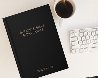 May include: A black notebook with the text "BUDGETS, BILLS, & BIG GOALS" and "KATE'S NOTES" in gold lettering. The notebook is on a white surface with a white keyboard and a cup of tea.