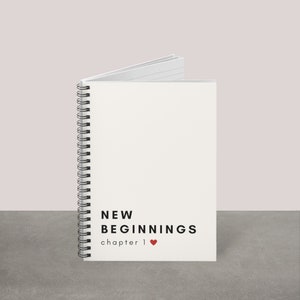 May include: A white spiral-bound notebook with the words "NEW BEGINNINGS chapter 1" printed in black. A small red heart is next to "chapter 1". The notebook is open, revealing lined pages.