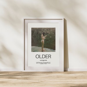 Peut inclure: Une affiche encadrée blanche avec le titre "OLDER" de Lizzy McAlpine. L'affiche présente une photographie d'une personne debout sur un promontoire rocheux surplombant une rivière et une forêt.