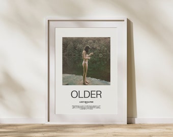 May include: A white framed poster with the title "OLDER" by Lizzy McAlpine. The poster features a photograph of a person standing on a rock ledge overlooking a river and forest.