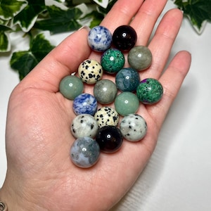 May include: A collection of 15 smooth, polished gemstone spheres in various colors, including green, blue, black, and white. The spheres are displayed in a hand.