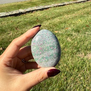 May include: A smooth, oval-shaped stone with a speckled pattern of gray, green, and pink hues. The stone is held in a hand against a backdrop of green grass and a sunny outdoor setting.