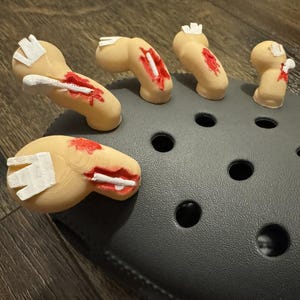 May include: A dark gray foam clog shoe featuring five beige, three-dimensional finger-shaped charms attached to the top. Each finger charm displays a simulated injury with red paint and a white bone-like protrusion, creating a textured, gory effect.