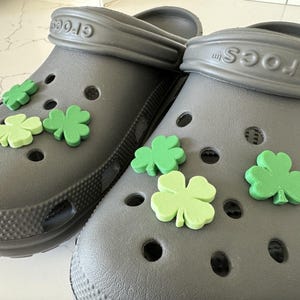 Op de afbeelding: Een paar grijze Crocs-klompen versierd met groene klaverbladbedels. De klompen hebben meerdere ventilatiegaten en een gestructureerde zool. De klavers zijn in verschillende groentinten.