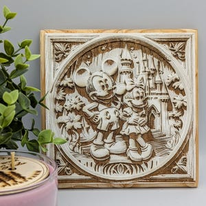 May include: A square, wooden plaque with a carved relief of Mickey and Minnie Mouse in front of a castle. The artwork is in a circular frame with floral and decorative elements. The plaque is light brown with white accents.