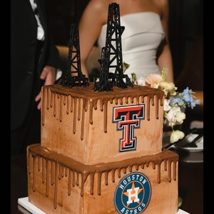 Puede incluir: Un pastel de boda de dos niveles decorado con glaseado de chocolate y un efecto de goteo. El nivel superior presenta un logotipo "T" rojo y el nivel inferior presenta un c&iacute;rculo azul con "HOUSTON ASTROS" y una "H" blanca en su interior. El pastel est&aacute; coronado con dos torres de perforaci&oacute;n de petr&oacute;leo negras.