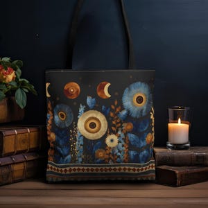 May include: A black tote bag featuring a printed design of blue and gold flowers, celestial moon phases, and a geometric border pattern at the bottom. The bag has two black shoulder straps and a rectangular shape with a dark, moody color palette.