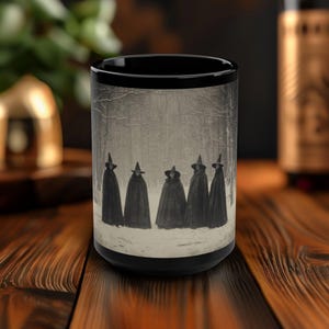 May include: A black coffee mug with a white image of six figures in black cloaks and pointed hats standing in a snowy forest. The figures are facing forward and appear to be walking in a line.