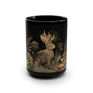 May include: A black ceramic mug featuring a detailed illustration of a brown jackalope with golden antlers, surrounded by golden botanical elements, mushrooms, and small stars against a dark background, creating a celestial forest scene.