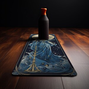 Puede incluir: Una botella negra cil&iacute;ndrica y alta con un tap&oacute;n naranja brillante se asienta sobre una alfombrilla rectangular. La alfombrilla presenta una ilustraci&oacute;n detallada de una figura con un vestido azul fluido que sostiene una balanza dorada.