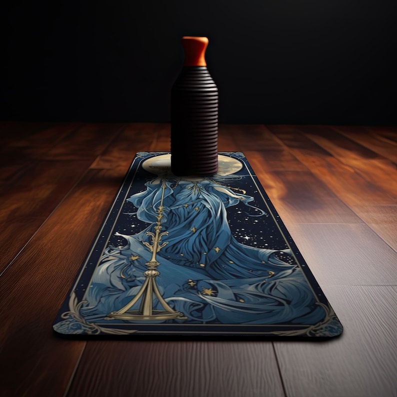 Puede incluir: Una botella negra cil&iacute;ndrica y alta con un tap&oacute;n naranja brillante se asienta sobre una alfombrilla rectangular. La alfombrilla presenta una ilustraci&oacute;n detallada de una figura con un vestido azul fluido que sostiene una balanza dorada.