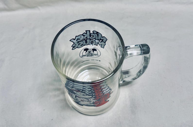 May include: Clear glass mug with a handle, featuring the text "Mickey Mouse" in a stylized font. The mug has a ribbed design and a graphic of Mickey Mouse. The design is in black, red, and white.