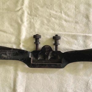 May include: A black, vintage-style drawknife with two adjustment screws and a curved blade. The tool has two handles with holes for hanging. The drawknife is designed for woodworking and shaping materials.