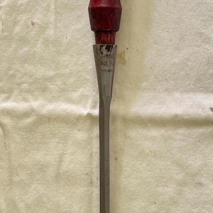 May include: A chisel with a dark red wooden handle and a silver-colored blade. The handle shows signs of wear, and the blade is rectangular. The tool is designed for woodworking and general construction.