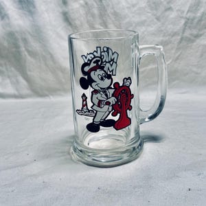 May include: Clear glass beer mug featuring a cartoon Mickey Mouse design. Mickey is dressed as a captain, holding a red ship's wheel. The mug has a handle and the words "Mickey Mouse" are printed above the character.