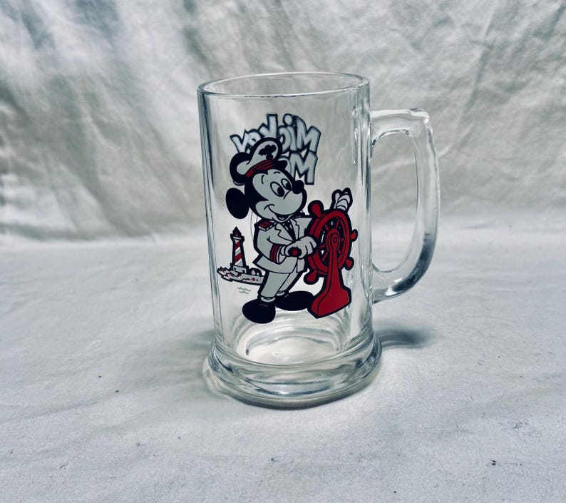 May include: Clear glass beer mug featuring a cartoon Mickey Mouse design. Mickey is dressed as a captain, holding a red ship's wheel. The mug has a handle and the words "Mickey Mouse" are printed above the character.