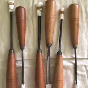 May include: A collection of woodworking chisels with wooden handles and metal blades. The chisels vary in size and shape, with some having straight blades and others having angled blades. The handles are a light brown color, and the blades are silver.