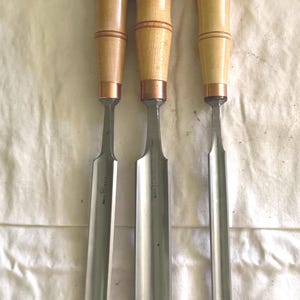 May include: Three wood chisels with light-colored wooden handles and copper-colored ferrules. Each metal blade features a Buck Brothers stamp and is protected at the tip by blue painter's tape and a small piece of tan paper with handwritten markings.
