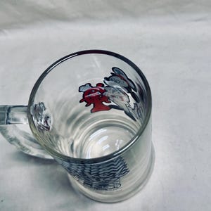 May include: Clear glass beer mug with a handle. The mug features a design of cartoon characters in red, white, and gray. The bottom of the mug has a decorative pattern. The mug is empty.