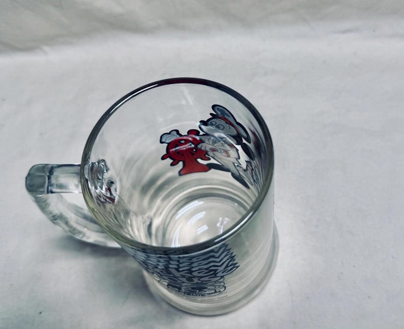 May include: Clear glass beer mug with a handle. The mug features a design of cartoon characters in red, white, and gray. The bottom of the mug has a decorative pattern. The mug is empty.