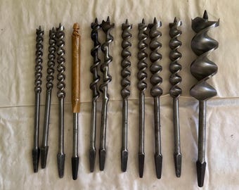 May include: A collection of vintage drill bits, including auger bits and twist drill bits, arranged on a light-colored surface. The bits vary in size and design, with a mix of spiral and straight fluted styles. One bit has a wooden handle.
