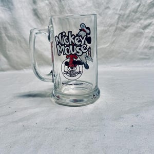 May include: Clear glass mug featuring a Mickey Mouse graphic. The mug has a handle and a rectangular shape. The Mickey Mouse graphic includes the words "Mickey Mouse" and an image of Mickey Mouse.