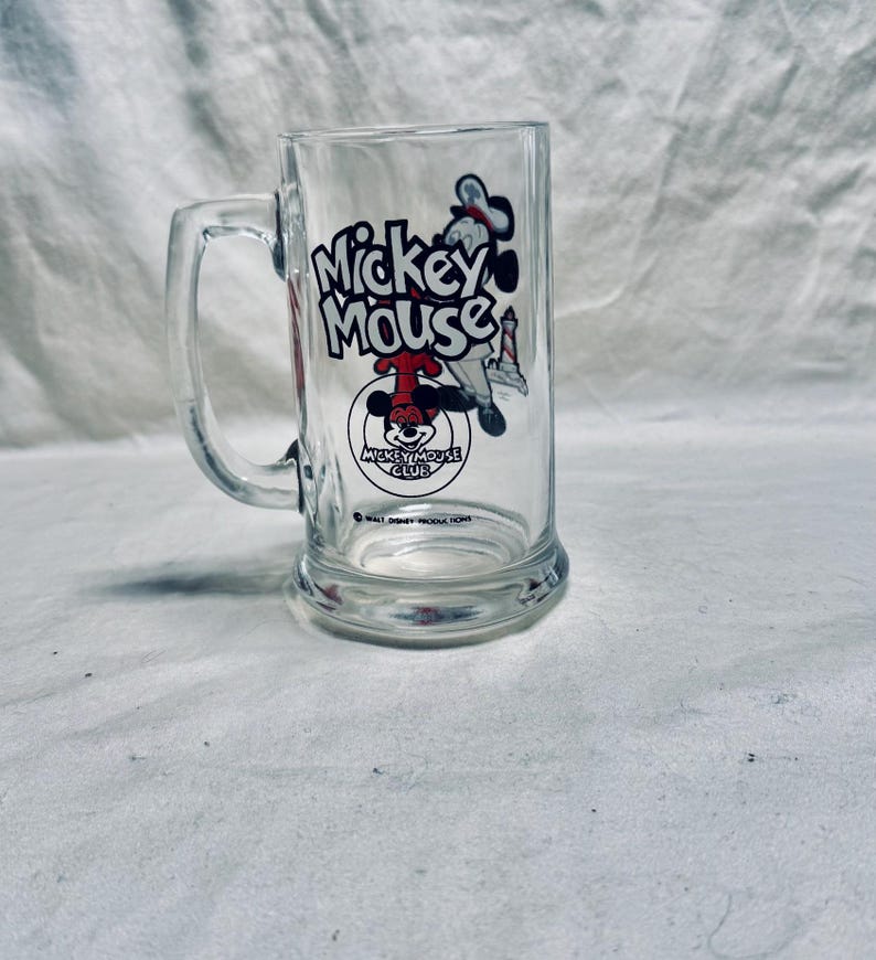 May include: Clear glass mug featuring a Mickey Mouse graphic. The mug has a handle and a rectangular shape. The Mickey Mouse graphic includes the words "Mickey Mouse" and an image of Mickey Mouse.