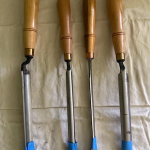 May include: Four woodturning chisels with light-colored wooden handles and metal ferrules. Each tool features a long steel blade, with three of the blades having blue painter's tape applied to the tips and one having tape on the handle end.