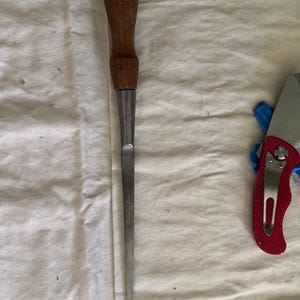 May include: A woodworking chisel with a wooden handle and a metal blade. The handle is a dark brown color, and the blade is silver. The chisel is lying on a white cloth.