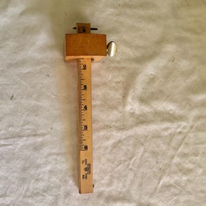 May include: A wooden marking gauge with a rectangular head and a graduated ruler. The ruler is marked with inch measurements. A metal thumbscrew is visible on the side of the head. The tool is used for woodworking.