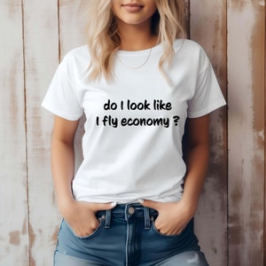 Pode incluir: Camiseta branca com o texto "do I look like I fly economy?" em preto. A camisa é usada com shorts jeans azuis. A modelo está em frente a uma parede de madeira marrom claro.