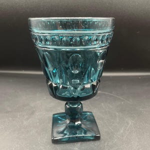 May include: A teal-colored, vintage-style goblet with a square base and a detailed, patterned bowl. The glass features a decorative design around the rim and on the body, adding a touch of elegance to the glassware.