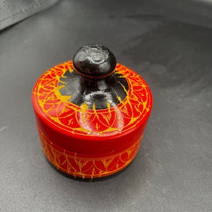 Könnte beinhalten: Eine kleine, runde, rote Holzkiste mit einem schwarzen Deckel. Die Kiste ist mit goldenen Mustern verziert.