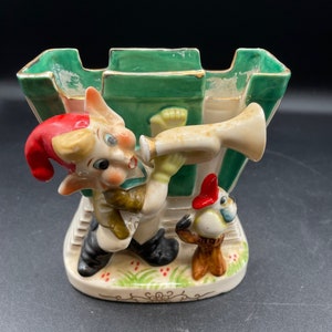 Op de afbeelding: Een keramiek beeldje van een elf die trompet speelt, met een kleine vogel die op een boomstronk zit. De elf draagt een rode muts, een groen shirt en een bruine broek. Het beeldje is geschilderd in felle kleuren en heeft een fantasierijk ontwerp.