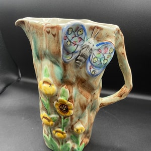 May include: A ceramic pitcher shaped like a tree trunk with a butterfly and yellow flowers painted on it. The pitcher has a handle and a spout.