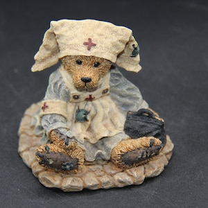 Op de afbeelding: Een klein, bruin teddybeerfiguurtje gekleed als verpleegster. De beer draagt een witte verpleegstersmuts met een rood kruis, een witte jurk met een rood kruis en een blauw jasje. De beer houdt een klein zwart tasje vast. De beer zit op een stenen basis.