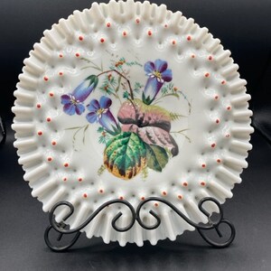 Pode incluir: Um prato de porcelana branca com uma borda festonada e um desenho floral. O prato tem uma borda elevada com pequenos pontos vermelhos ao redor da borda. O desenho floral apresenta flores azuis, folhas verdes e uma flor rosa.