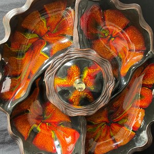 May include: A black ceramic serving dish with four sections. The dish has a swirling pattern of orange, red, and yellow glaze. The centre section has a raised, circular design with a gold knob.