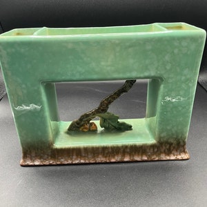 May include: A green ceramic planter with a rectangular shape and a cut-out design featuring a branch with leaves and acorns. The planter has a brown glaze on the bottom edge.